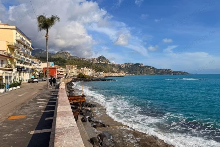 Wellen brechen an schwarzen Steinen vor einer Straße am Meer